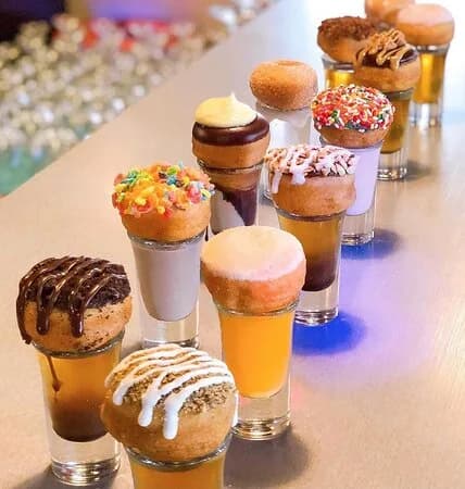 Donut Distillery mini donuts with assorted toppings balanced on filled shot glasses lined up on a bar