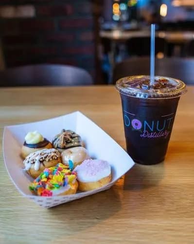 Donut Distillery paper tray of six topped mini donuts next to an iced coffee in a branded cup