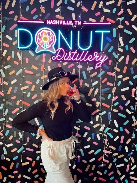 Guest sipping a drink in front of the neon Donut Distillery Nashville, TN sign on a sprinkle-patterned wall