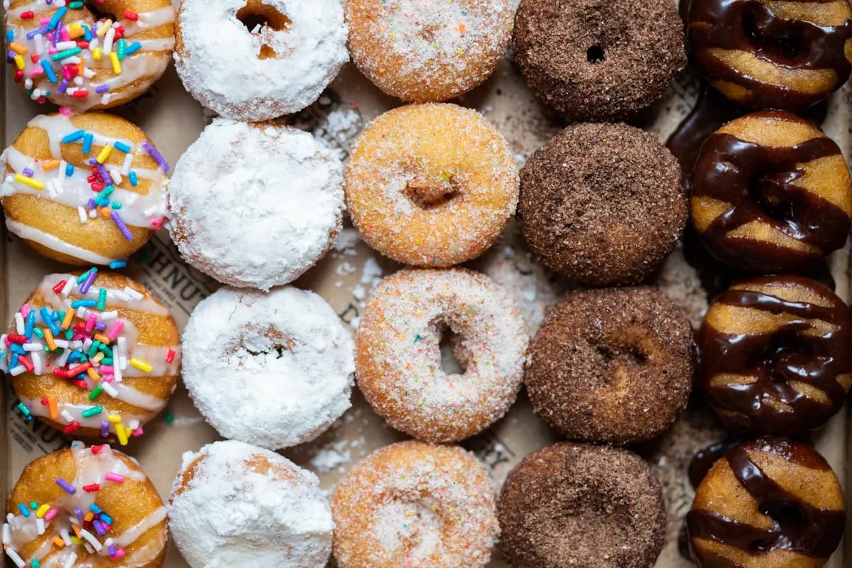 Overhead view of Doughnuttery mini doughnuts in rows: rainbow sprinkle, powdered sugar, flavored sugar, cinnamon sugar, and chocolate drizzle
