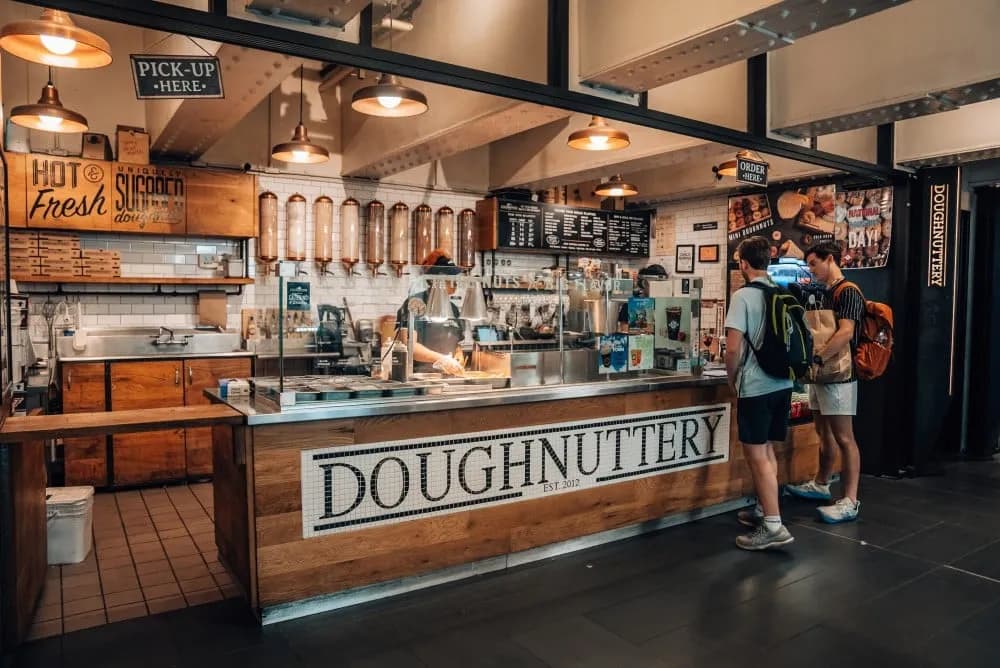 Doughnuttery counter in Chelsea Market with a tiled Doughnuttery sign, sugar dispensers on the back wall, and customers ordering