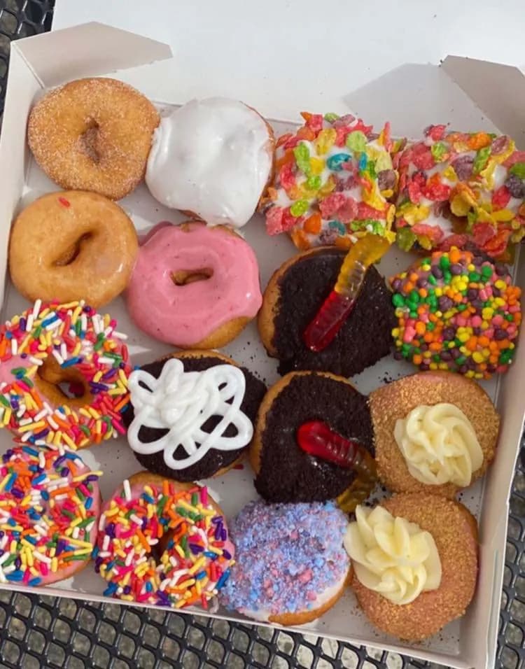 Box of assorted Niky's mini donuts with cinnamon sugar, pink icing, rainbow sprinkles, fruity cereal, cookie crumbs with gummy worms, and cream swirls