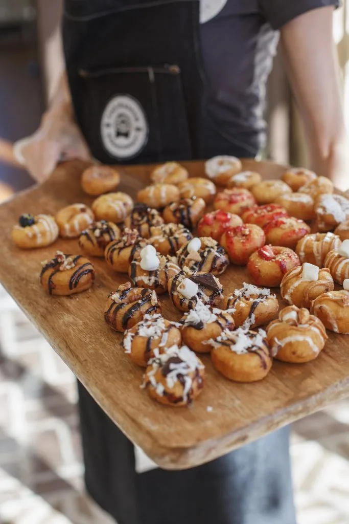 Server in a Dapper Doughnut apron holding a wooden board of mini doughnuts with chocolate drizzle, marshmallows, and red glaze