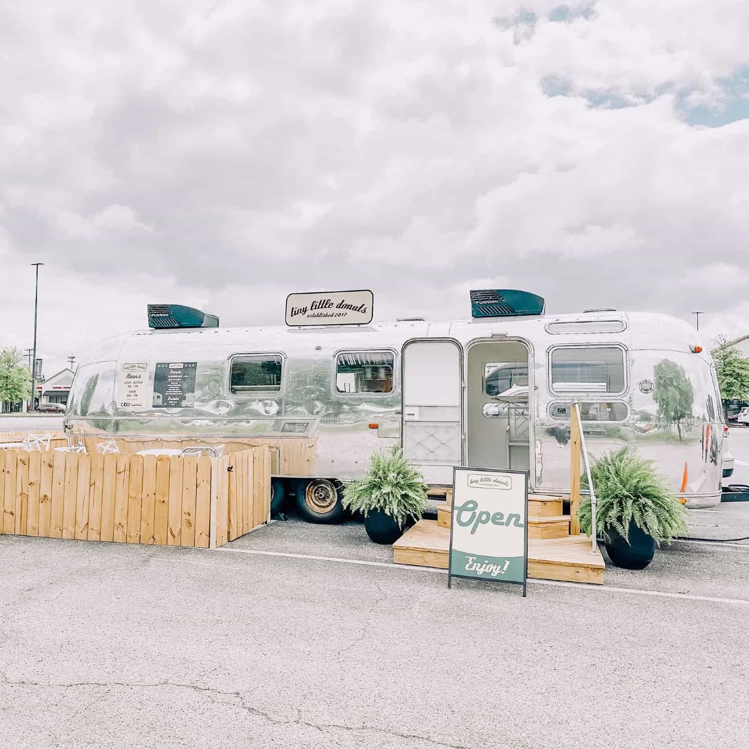 Tiny Little Donuts silver vintage Airstream trailer with an Open sign, wooden deck, and potted ferns in a parking lot