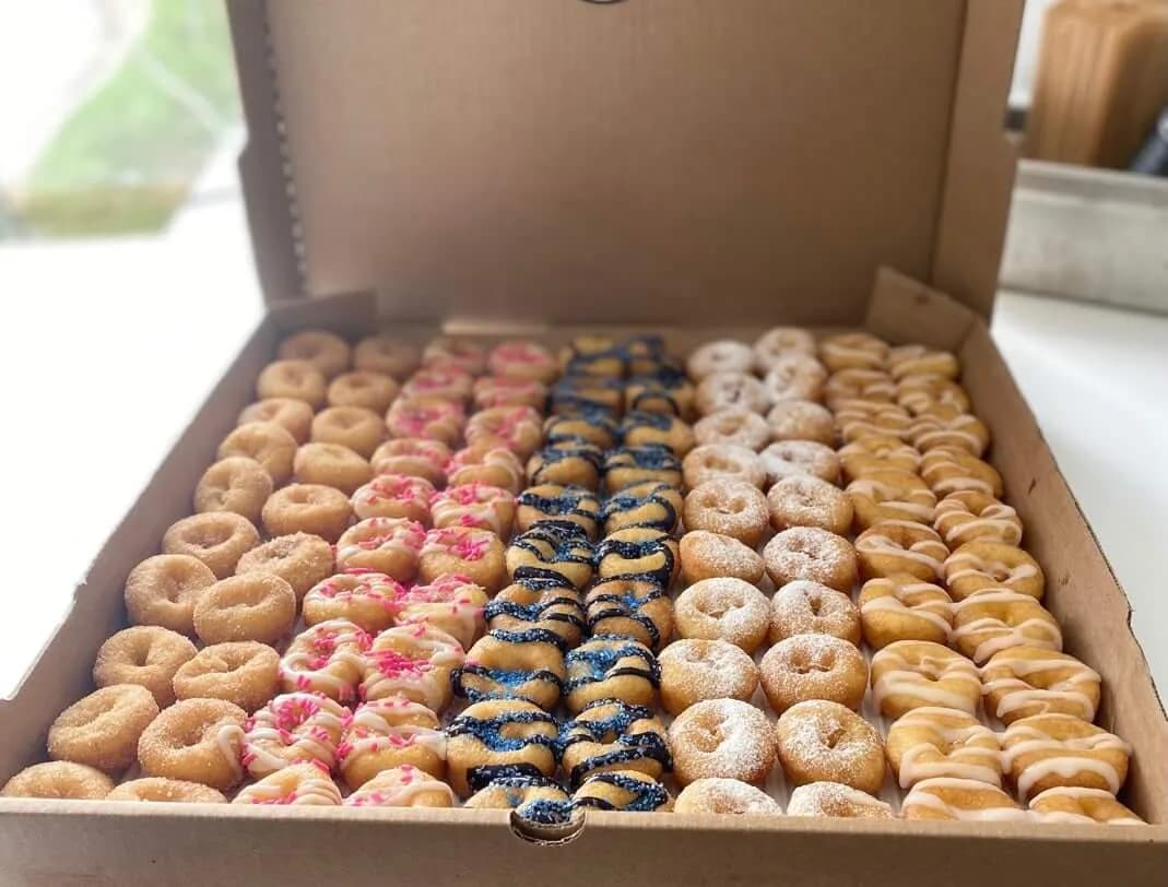 Large open box of Tiny Little Donuts mini donuts in rows of cinnamon sugar, pink drizzle, blue drizzle, powdered sugar, and white icing