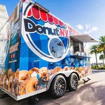 Blue and red DonutNV mini donut trailer with a serving window and round viewing window, parked outdoors near palm trees