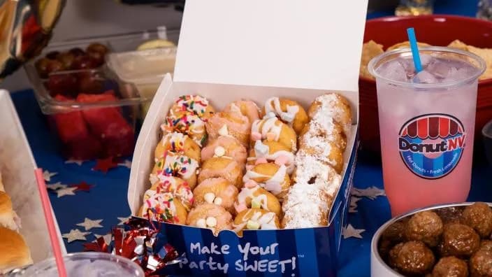 Open DonutNV box of sprinkle, drizzled, and powdered-sugar mini donuts beside a DonutNV lemonade on a party table